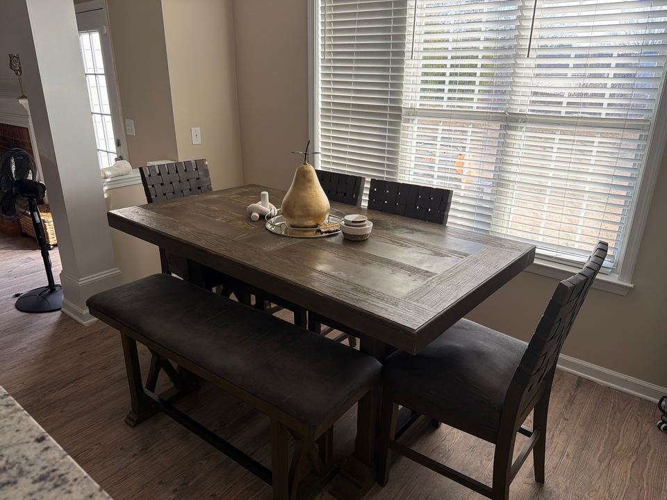 Dining area with a dark wood farmhouse table, an upholstered bench and chairs beside a large window with white blinds