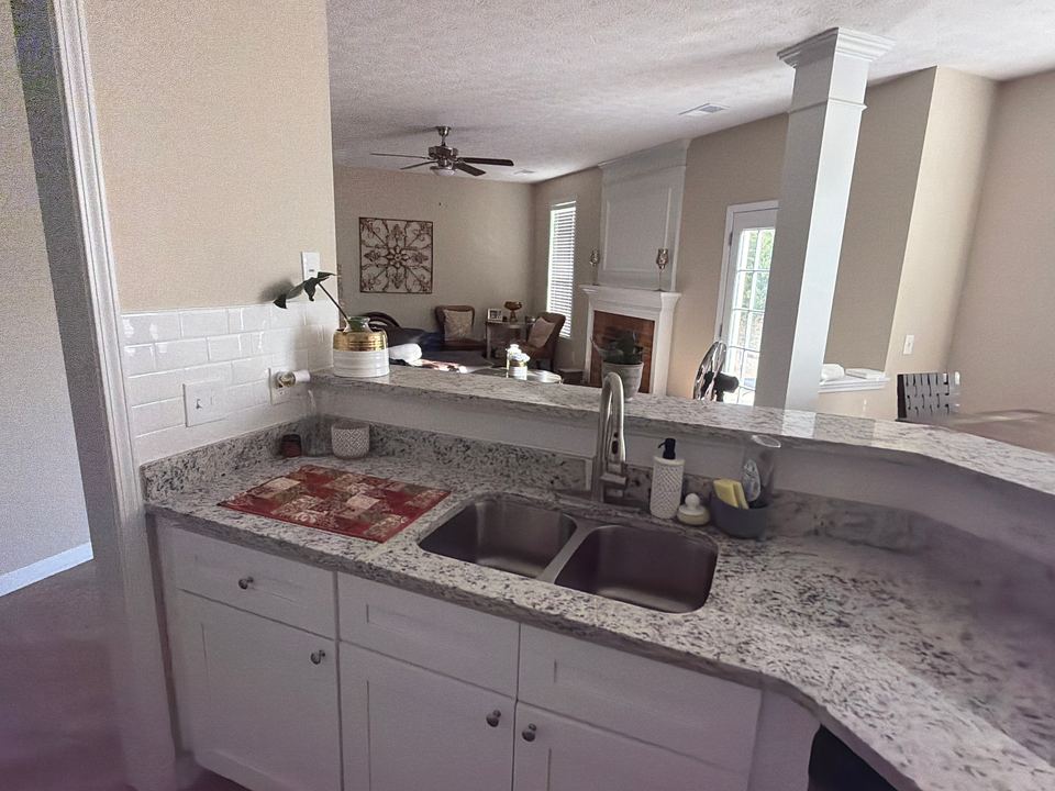 White kitchen with speckled granite countertops, a double stainless-steel sink and white cabinets, opening onto a living room