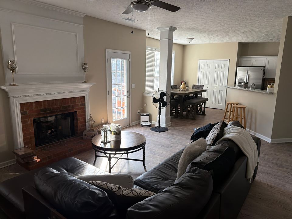 Open living room with a brick fireplace, a black leather sectional and a dining table leading to the kitchen